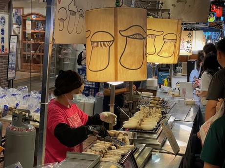 A vendor in a pink shirt and mask preparing mushroom skewers under hanging lanterns with mushroom illustrations, in a bustling food market.