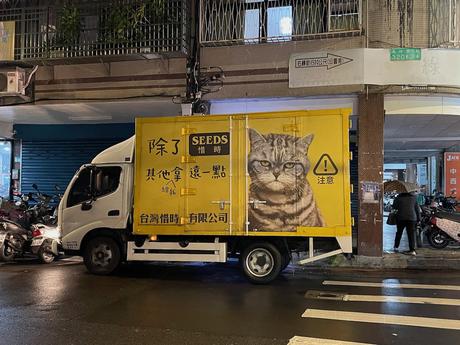 A parked yellow truck featuring a graphic of a grumpy-looking cat and text, parked on a rainy street with scooters nearby.