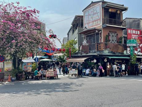 A lively street food market with tables and people under a blooming bougainvillea tree, with shops and hanging decorations along the street.