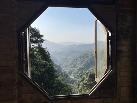 A scenic view through an open octagonal window framing a forested valley under a bright blue sky.