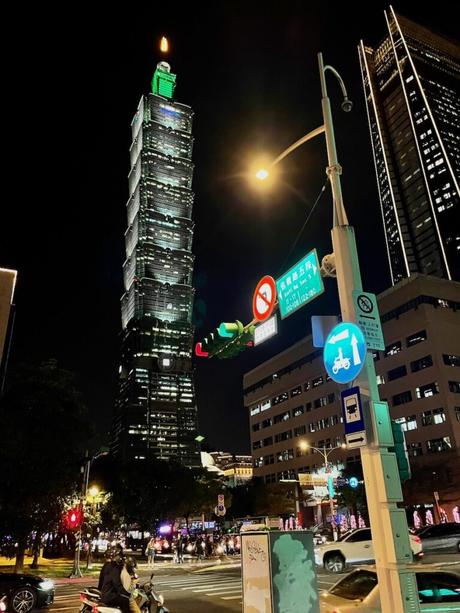 Taipei 101 towering above a busy intersection at night, with streetlights, traffic, and signs creating a lively urban scene.