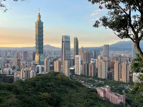 A scenic sunset view of Taipei's skyline with Taipei 101 towering prominently amidst the city, surrounded by lush greenery and golden light.