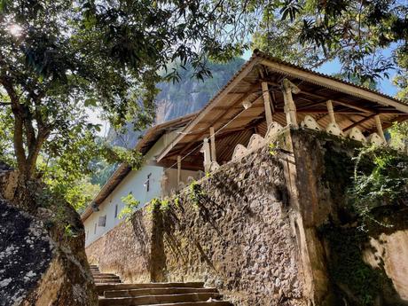 A stone staircase leading up to a traditional temple building nestled among trees, with sunlight filtering through the foliage.