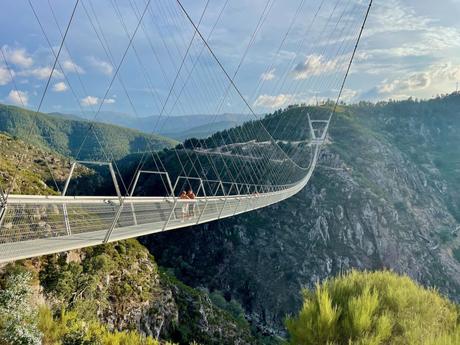 Two people stand in the middle of the 516 Arouca suspension bridge that spans across a canyon, with a view of the green mountains and forests in the distance. The bridge appears delicate against the vast landscape.