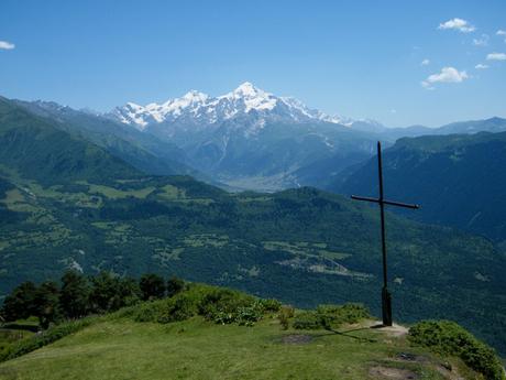 A panoramic view of the Caucasus mountain range in Georgia with a snowy peak in the center. A tall black cross stands on a grassy hill in the foreground overlooking the valley.