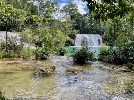 A scenic view of two waterfalls cascading into a turquoise pool surrounded by dense greenery and rocks.