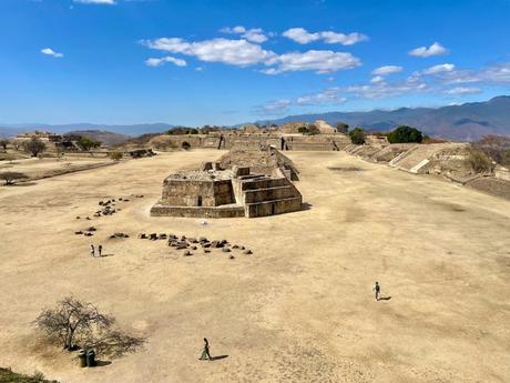 A wide view of ancient ruins at Monte Albán, with large stone structures under a bright blue sky and mountains in the distance.