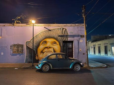 A street at night with a mural of an elderly woman's face painted on a lavender wall, with an old Volkswagen Beetle parked in front and a streetlight illuminating the scene.