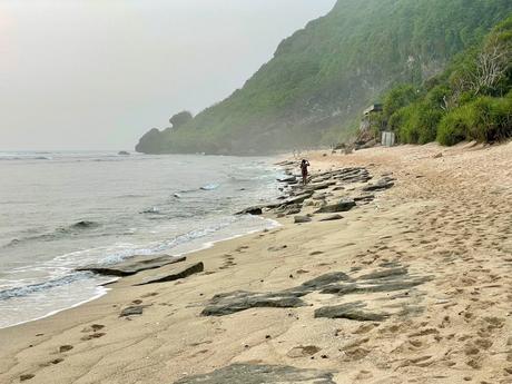 Golden sands of Nyang Nyang Beach, Uluwatu, with sea spray in the air, a person walking on the beach in the distance, and lush green cliffs in the background