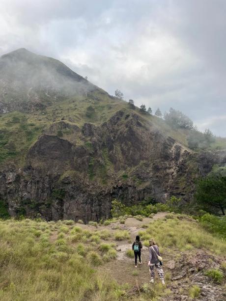 Two people hiking up a mist-covered grassy hill with rocky outcroppings, surrounded by lush greenery.