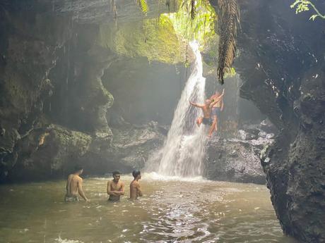 A group of young men playfully swim near a small waterfall in a cave, with one of them mid-jump from the waterfall.