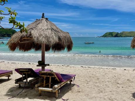 Two bamboo beach loungers with thatched umbrellas on a white sand beach, facing the calm turquoise waters, with a fishing boat and green hills in the background.