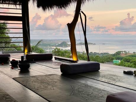 A serene yoga studio overlooking the coastline of Kuta, Lombok at sunset, with mats and bowls arranged on a wooden platform that opens up to a panoramic view of the ocean and distant islands.