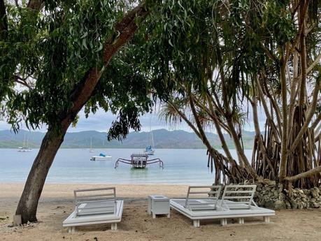 Two white lounge chairs under a tree facing the water, with boats anchored offshore. Large branches create a shaded area near the calm coastline.