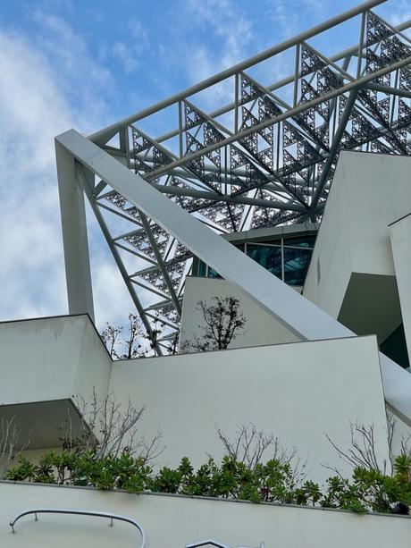 A modern architectural building featuring white angular structures and a triangular metal framework, set against a blue sky with scattered clouds.