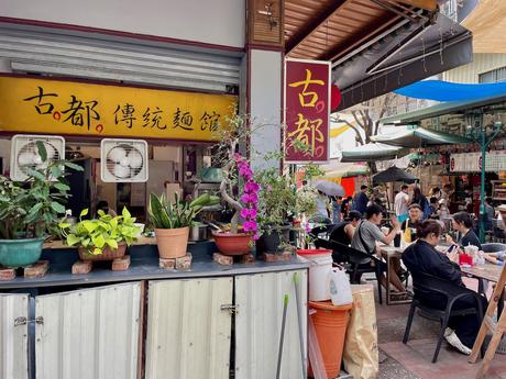 A bustling street-side noodle shop with yellow signage, potted plants, and a crowd of people enjoying their meals at outdoor tables under a canopy.