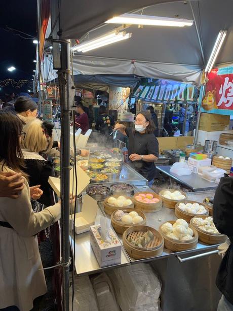 A lively night market food stall selling steamed buns, dumplings, and colorful desserts, with a vendor serving customers amidst steaming trays.