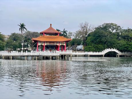 A serene lakeside pavilion with a traditional red roof, surrounded by lush greenery and a white arched bridge, reflected in the calm water.