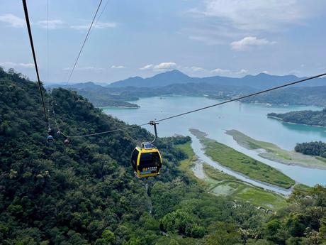 A cable car suspended over lush green forests and the sparkling blue waters of Sun Moon Lake, with distant mountains forming a scenic backdrop.