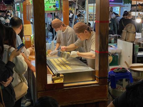 Two masked vendors pouring batter onto a griddle to make small round desserts, surrounded by customers at a popular night market stall.