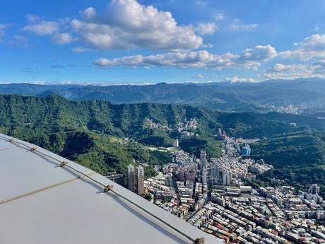 A sweeping view of green mountains and cityscape as seen from Taipei 101, under a vivid blue sky with fluffy clouds.