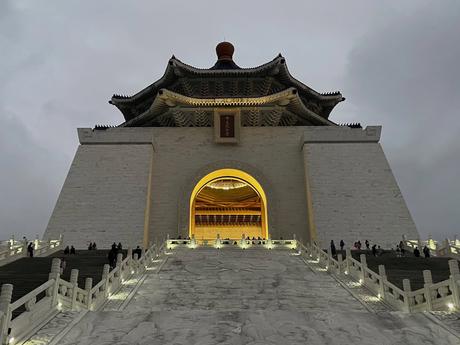 The Chiang Kai-shek Memorial Hall in Taipei illuminated at dusk, with a grand white staircase leading up to the archway and its iconic octagonal roof visible against the cloudy sky.