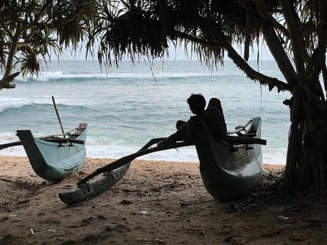 A fisherman resting in a traditional wooden canoe on the beach under the shade of a tree, with the ocean in the background.