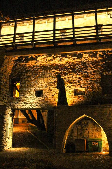 A medieval stone wall illuminated by yellow lights at night, with a statue of a hooded figure standing in a niche. An archway beneath the wall leads to a cobblestone path.