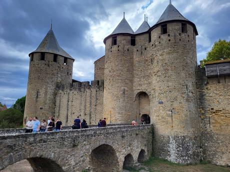 The medieval castle of Carcassonne, France, featuring tall stone towers and battlements under a cloudy sky. An arched stone bridge in the foreground leads to the castle entrance, with a group of people walking across.