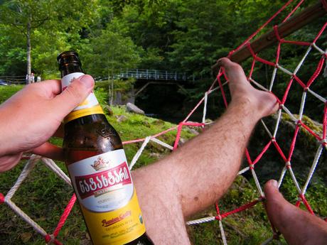 A relaxed scene in Georgia, with a point-of-view shot taken by Alex Tiffany lying in a red-and-white rope hammock holding a bottle of Georgian beer labeled in local script, with lush green trees and a small footbridge visible in the background.