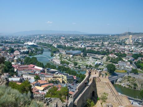 A view over Tbilisi with a river running through the center. A mix of modern and traditional architecture is visible, with hills and mountains in the background under a clear blue sky.