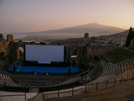 The ancient Greek theatre in Taormina, Sicily, set against a stunning backdrop of Mount Etna and the coastline. The theatre is prepared for a film festival, with rows of seating and a large screen on the stage.