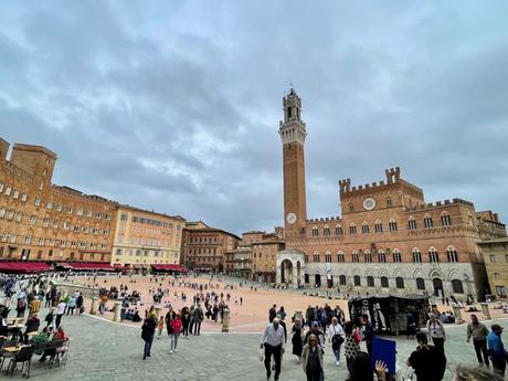 A wide-angle shot of Piazza del Campo in Siena, Italy, with crowds of people walking through the large square, surrounded by historical buildings and dominated by the tall Torre del Mangia under a cloudy sky.