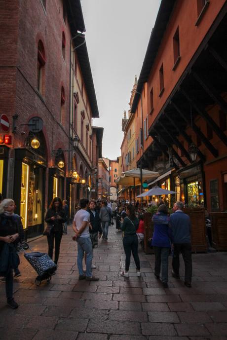 A lively street in the historical centre of Bologna, Italy, lined with historic red and orange buildings featuring arched windows and small shops. People walk and gather along the cobblestone street, while cafes with outdoor seating add a bustling atmosphere.