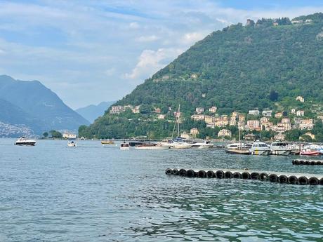 A peaceful scene on the shores of Lake Como in Italy with boats docked on the water and houses nestled along the hills, with green mountains rising in the distance.