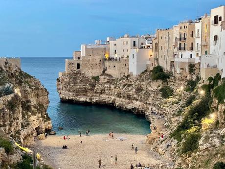 The picturesque beach at Polignano a Mare, Sicily, surrounded by dramatic limestone cliffs and historic white buildings perched above. The clear turquoise waters meet a sandy cove, with people relaxing and exploring the area.