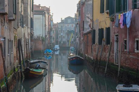 A tranquil, narrow canal with rustic boats docked along weathered buildings in Venice, with colorful laundry hanging on a line.