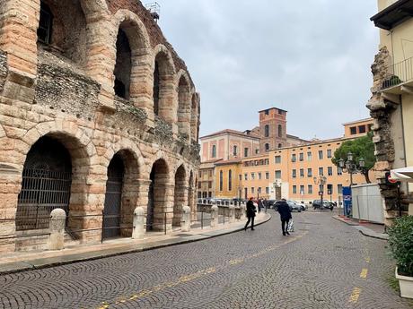 The iconic Verona amphitheater in Italy, showcasing its well-preserved Roman arches and stone structure. A quiet cobblestone street runs alongside, with modern buildings in the background.