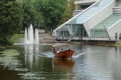 20 Best Places to Visit in Latvia A small wooden boat with a canopy cruises on a calm lake near a modern glass building, with water fountains and lush green trees in the background.