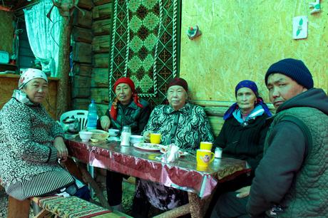 20 Best Places to Visit in Kyrgyzstan A group of older Kyrgyz people seated around a table inside a wooden house, enjoying a meal together. Their traditional headscarves and clothing add cultural depth to the cozy setting, while a woven tapestry decorates the wall.