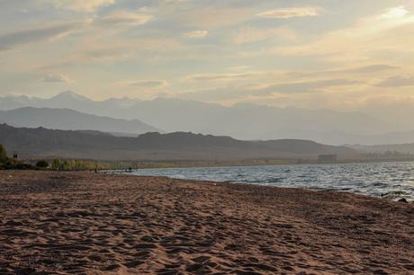 20 Best Places to Visit in Kyrgyzstan A peaceful lakeside view at sunset, with soft golden light reflecting off the water. The distant mountains create a layered backdrop, giving the image a sense of tranquility and natural beauty.
