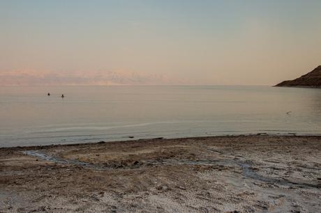 17 Best Places to Visit in Israel A tranquil view of the Dead Sea at sunset, with a calm shoreline in the foreground and two distant people floating in the water. The horizon shows a faint outline of mountains shrouded in a soft pink glow.