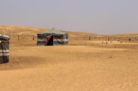 19 Best Places to Visit in Oman Two traditional Bedouin tents in the Omani desert, made of striped fabric, standing on the sandy ground with the arid, rolling dunes in the background.