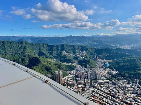 20 Best Places to Visit in Taiwan A sweeping view of green mountains and cityscape as seen from Taipei 101, under a vivid blue sky with fluffy clouds.