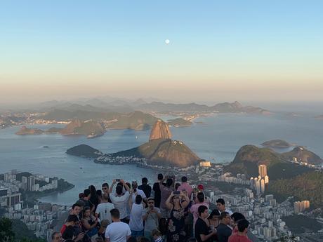 Sweeping view of Rio de Janeiro at sunset, taken from the top of the corcovado hill, with the city and islands laid out below. In the foreground there is a crowd of tourists taking photos.