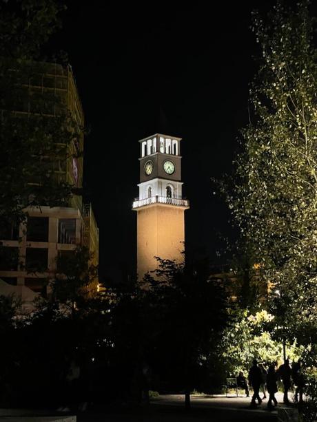 14 Best Things To Do In Tirana (& Surrounding Area) Tirana’s historic Clock Tower illuminated at night, standing tall against the dark sky. Trees and silhouettes of people walking in Skanderbeg Square add depth to the atmospheric scene.