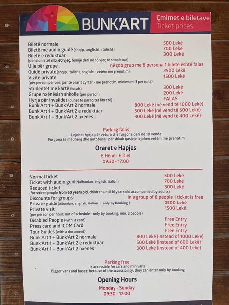 14 Best Things To Do In Tirana (& Surrounding Area) A signboard at the entrance of Bunk’Art, a museum in Tirana, Albania, displaying ticket prices, discounts, and opening hours in both Albanian and English. The colorful logo at the top contrasts with the detailed pricing information below.