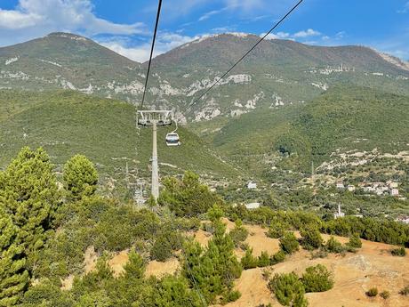 14 Best Things To Do In Tirana (& Surrounding Area) A scenic ride on the Dajti Ekspres cable car in Tirana, Albania, offering breathtaking views of the lush green mountains and scattered villages below. The gondola moves high above the trees under a clear blue sky.
