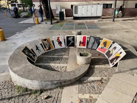 14 Best Things To Do In Tirana (& Surrounding Area) A small outdoor art display on a curved concrete structure in Tirana, Albania, showcasing modern, abstract paintings in black, red, and yellow tones. Two people are talking nearby, while a police officer stands in the background.