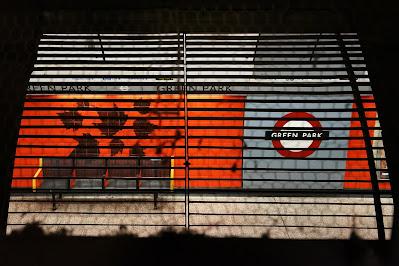 Hidden signs of Green Park station Through a grill, the wall of a platform is visible. It is orange and grey, with a 'Green Park' roundel and a row of four seats.